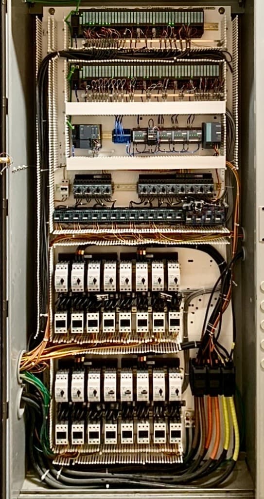 Industrial electrical control cabinet with PLC I/O modules, motor starters, organized wiring in cable duct, and power feeds.